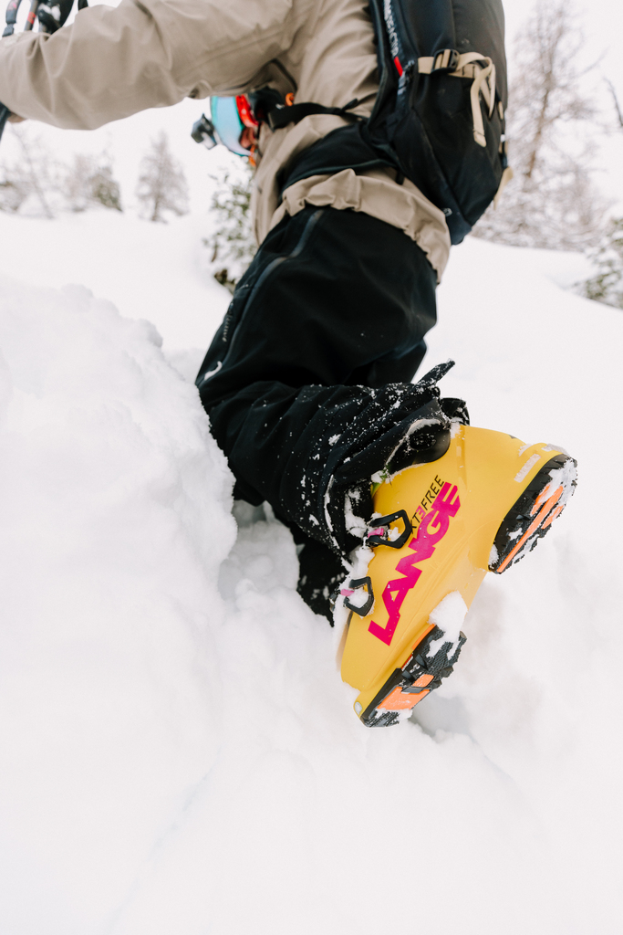 A close-up of a yellow Lange ski boot with pink lettering, walking through the snow.