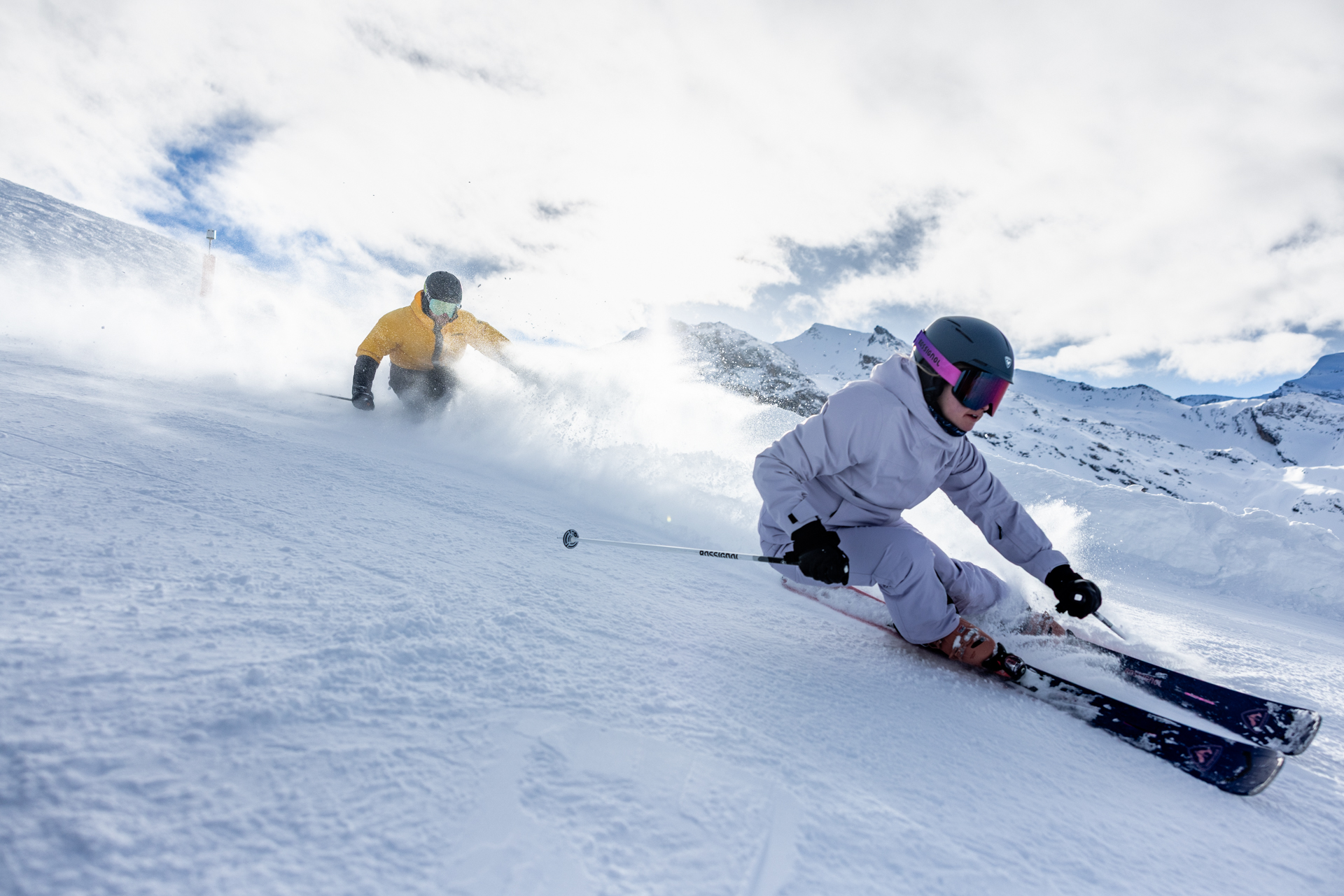 Two people skiing on the slopes. The person further up the slope is wearing purple, and the other is wearing yellow.